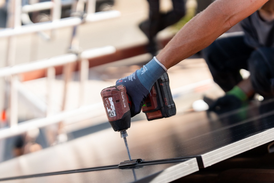 Eine Person installiert mit einer Bohrmaschine Solarmodule auf einem Dach und demonstriert die wesentlichen Schritte zum Verst&auml;ndnis der Funktion einer Photovoltaikanlage.