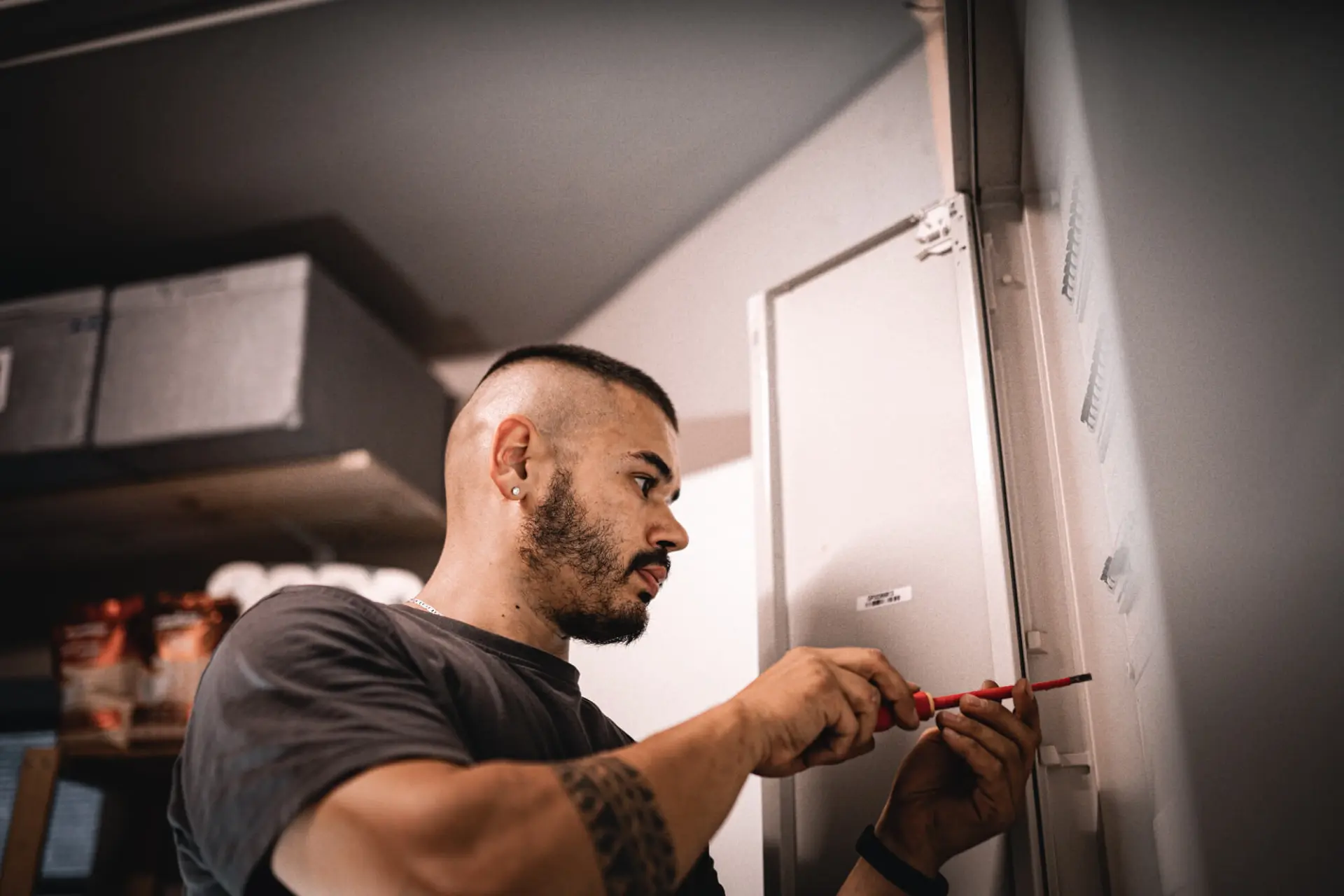 Ein b&auml;rtiger Mann arbeitet mit einem Schraubenzieher an einer Schalttafel in einem Raum und f&uuml;hrt Wartungsarbeiten an einem Smart Grid-System durch.