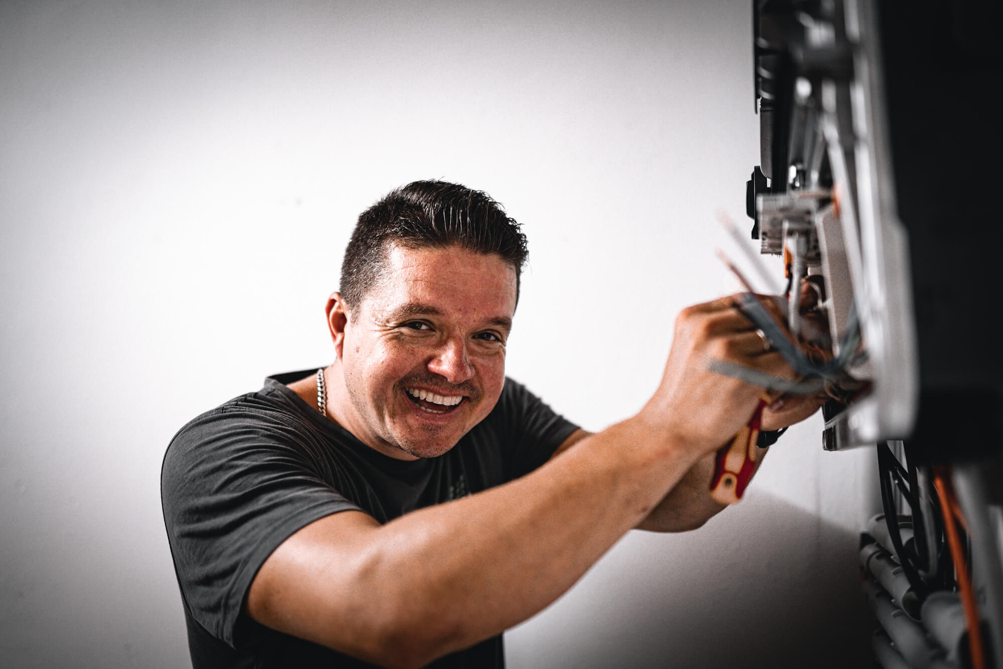 Ein l&auml;chelnder Mann arbeitet mit elektrischen Leitungen in der N&auml;he eines Stromz&auml;hlers und h&auml;lt ein Werkzeug vor einem wei&szlig;en Wandhintergrund.