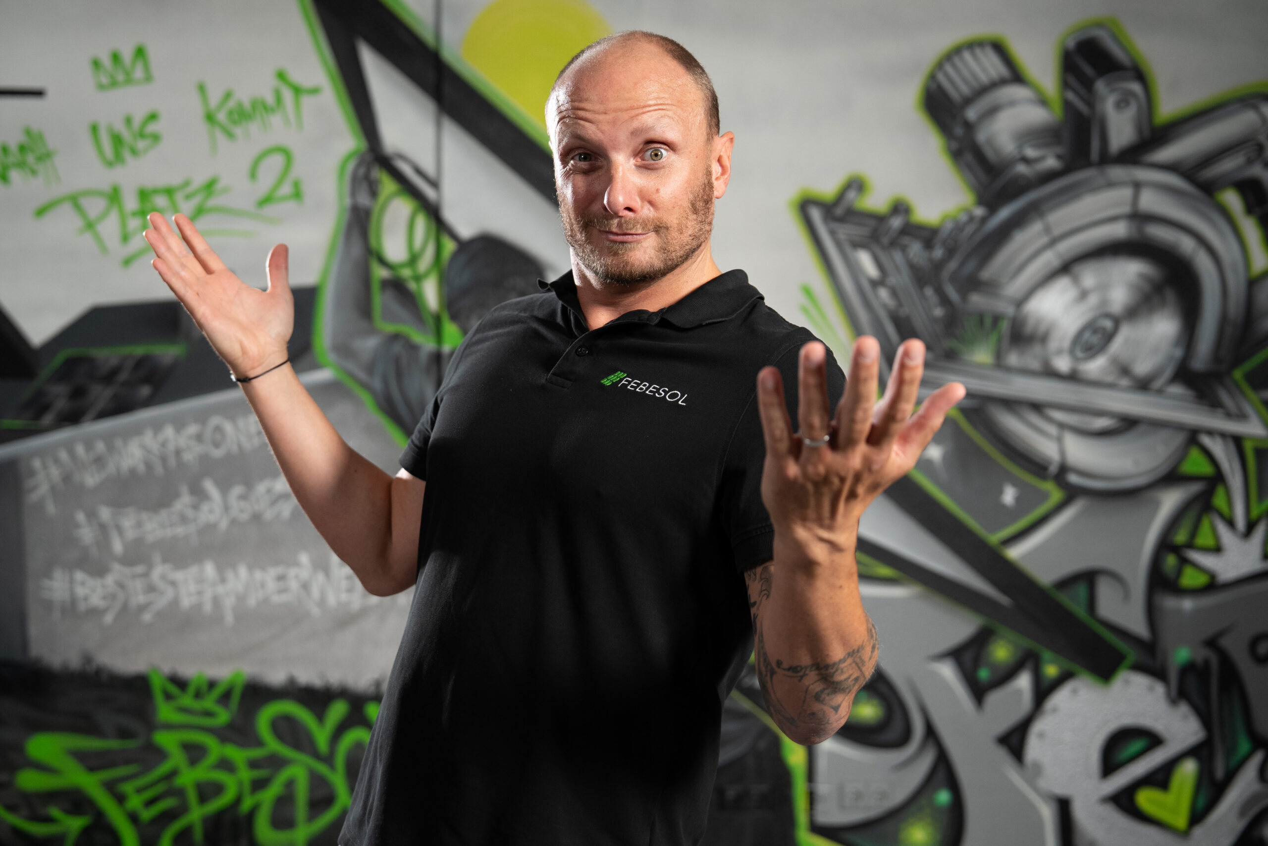 Ein Mann in einem schwarzen Polohemd zuckt mit einem verspielten Ausdruck vor einer bunten Graffiti-Wand.