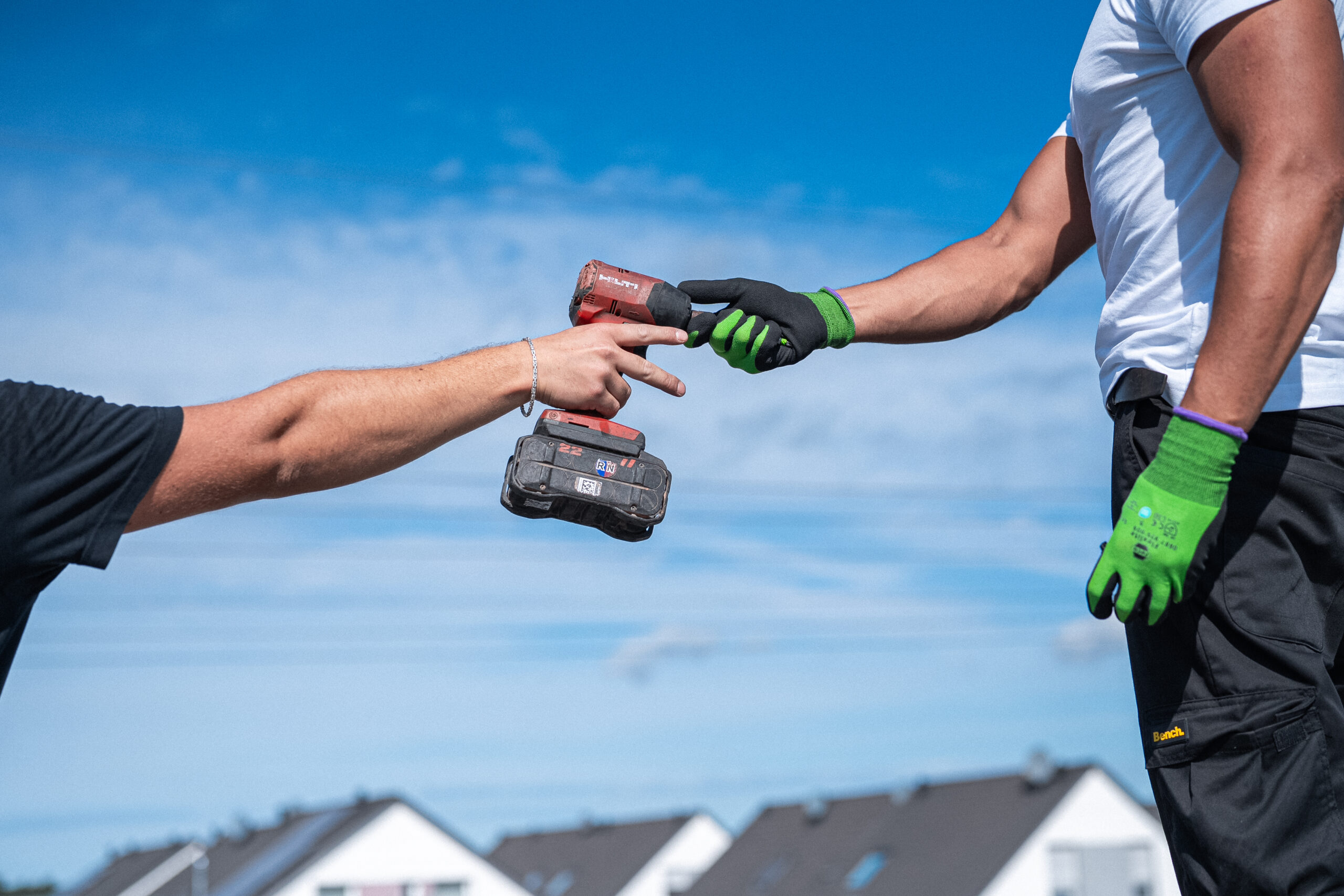 Zwei Arbeiter mit Handschuhen tauschen eine Bohrmaschine im Freien aus, mit H&auml;usern und blauem Himmel im Hintergrund.