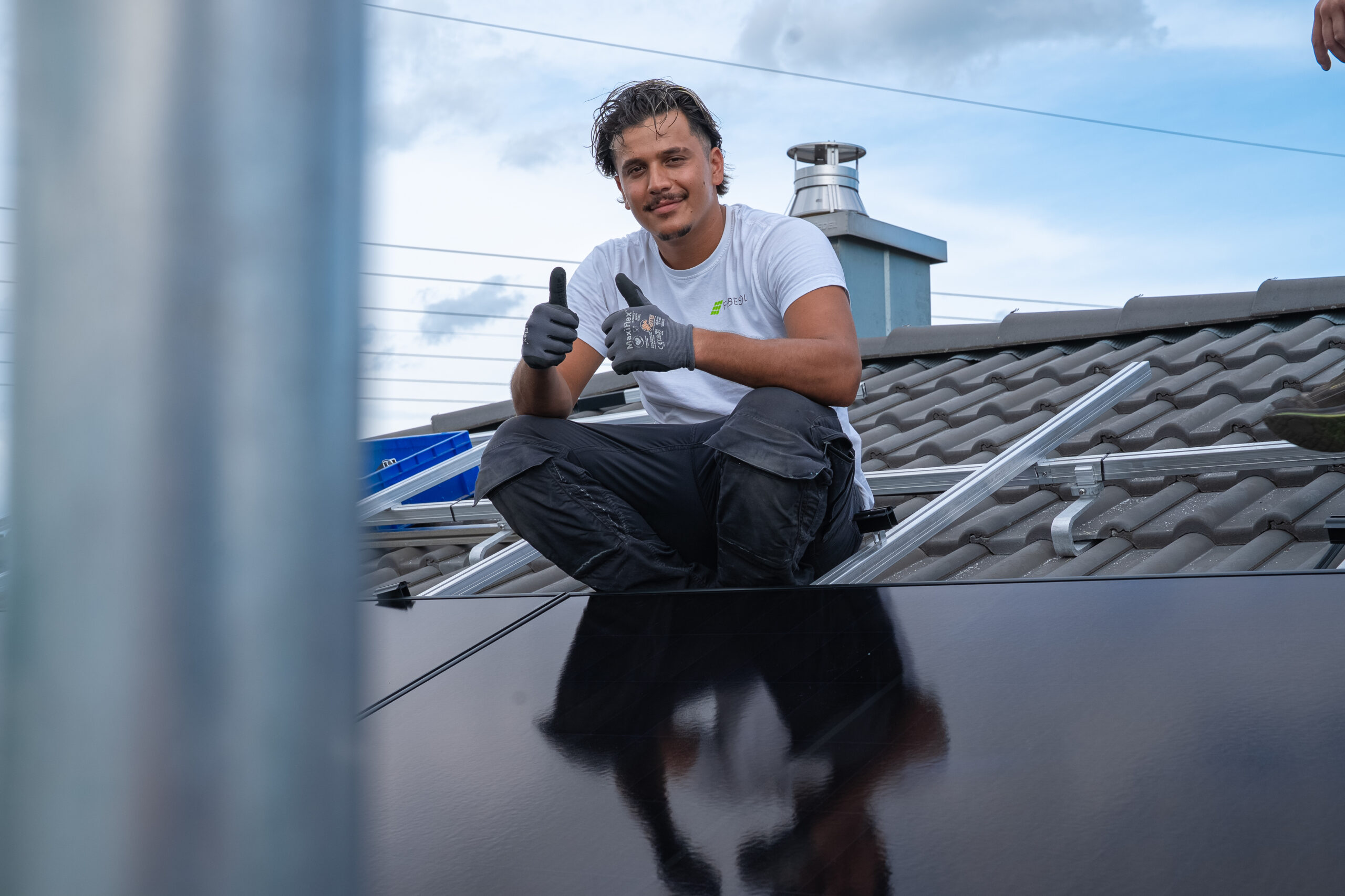 Mann sitzt auf einem Dach neben Solarzellen, l&auml;chelt und zeigt zwei Daumen nach oben, mit bew&ouml;lktem Himmel im Hintergrund.