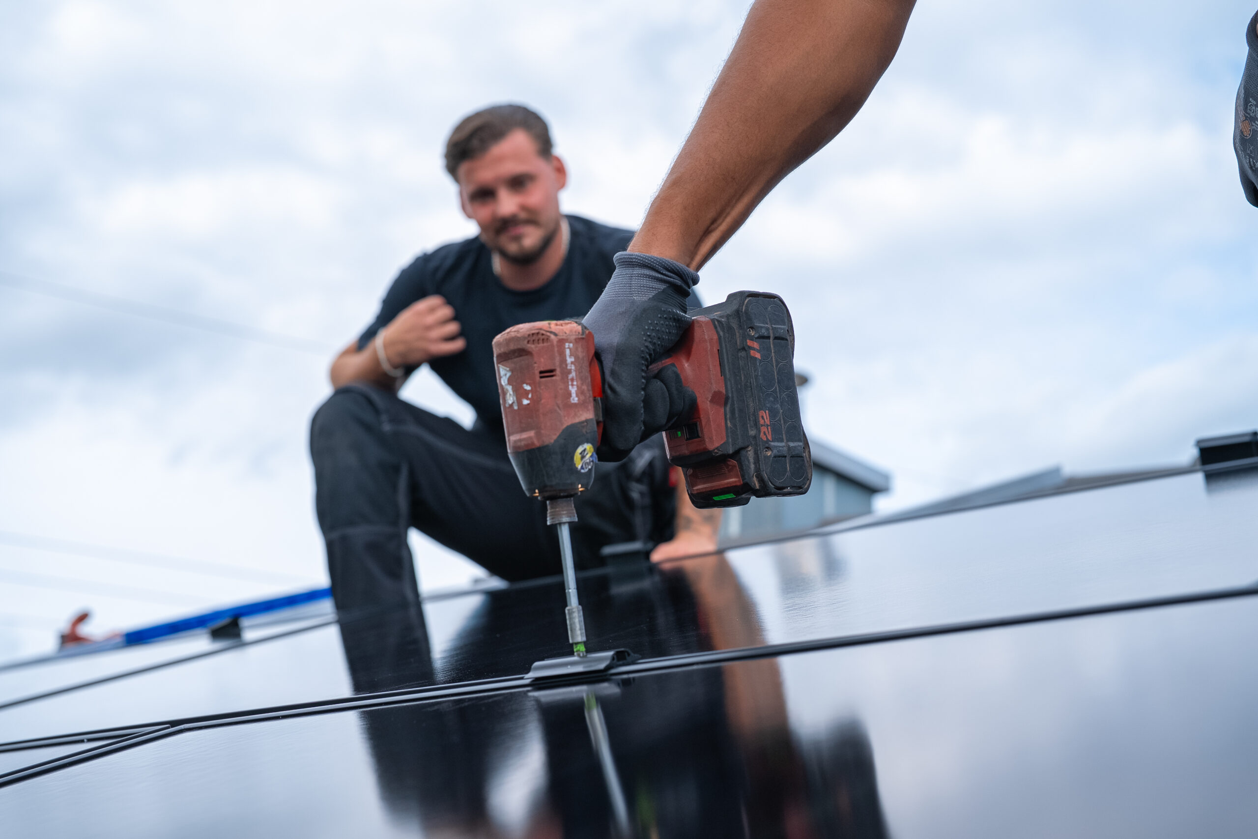 Ein Arbeiter installiert ein Solarmodul mit einer Bohrmaschine, w&auml;hrend eine andere Person im Hintergrund auf einem Dach kniet.