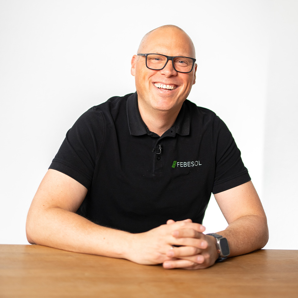 Ein lächelnder Mann mit Brille in einem schwarzen FEBESOL-Hemd sitzt an einem Holztisch vor einem weißen Hintergrund.