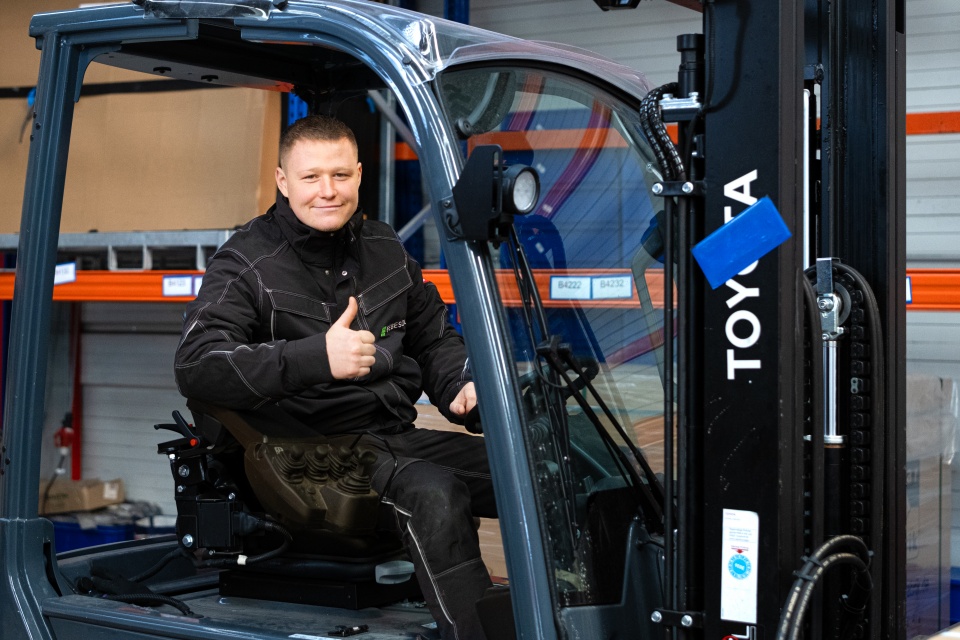 Ein Mann in Arbeitskleidung sitzt auf einem Gabelstapler, l&auml;chelt und zeigt den Daumen nach oben in einer Lagerhalle.