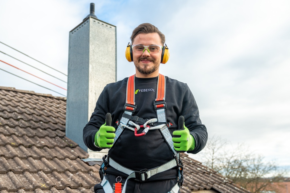 Ein l&auml;chelnder Arbeiter in Schutzkleidung steht auf einem Ziegeldach und zeigt zwei Daumen nach oben.