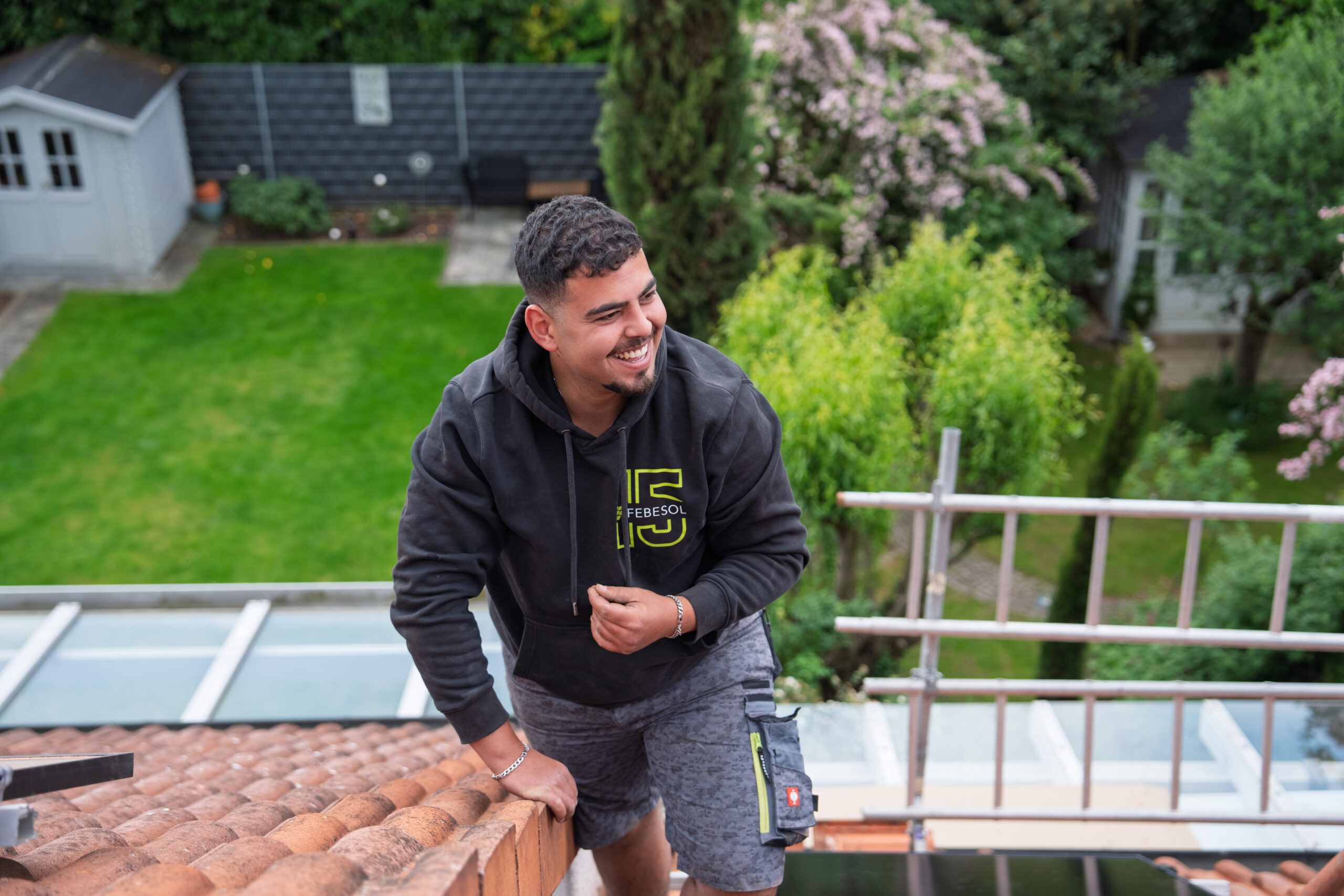 Ein l&auml;chelnder Mann in Arbeitskleidung steht auf einem Dach, im Hintergrund ist ein &uuml;ppiger Garten zu sehen.