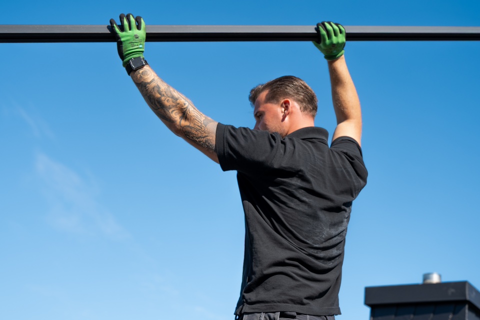 Ein Mann in schwarzem Hemd und gr&uuml;nen Handschuhen hebt eine Metallstange unter freiem Himmel an.